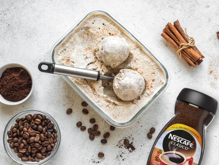Recipiente con helado y cuchara junto a astillas de canela, granos de café, café instantáneo y un frasco de Nescafé Clásico