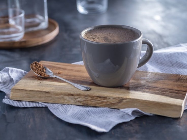Taza de chocolate color gris sobre una tabla y junto a esta una cuchara con chocolate en polvo y al fondo vasos de vidrio