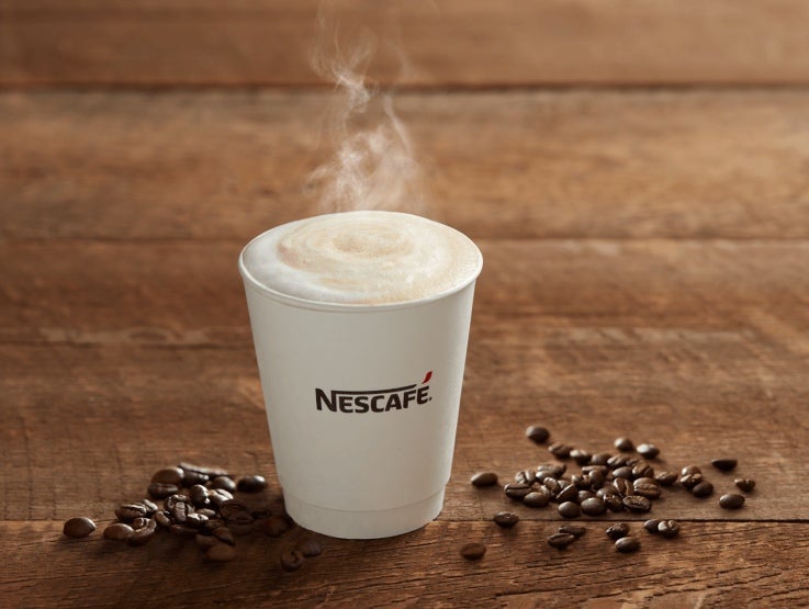 Vaso desechable blanco con el logo de NESCAFÉ® con una bebida caliente y alrededor granos de café tostados