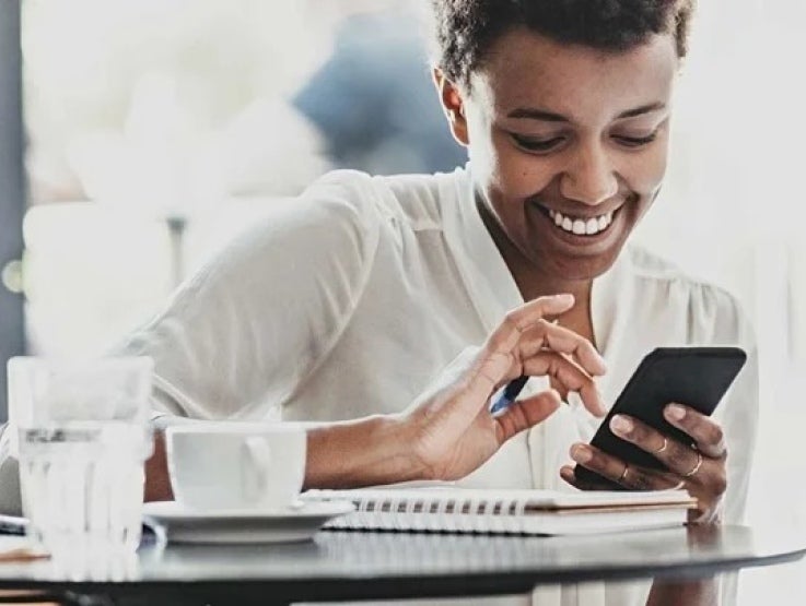 Mujer revisa su celular estando al frente de una mesa sobre la que hay una taza, un vaso de vidrio y un cuaderno