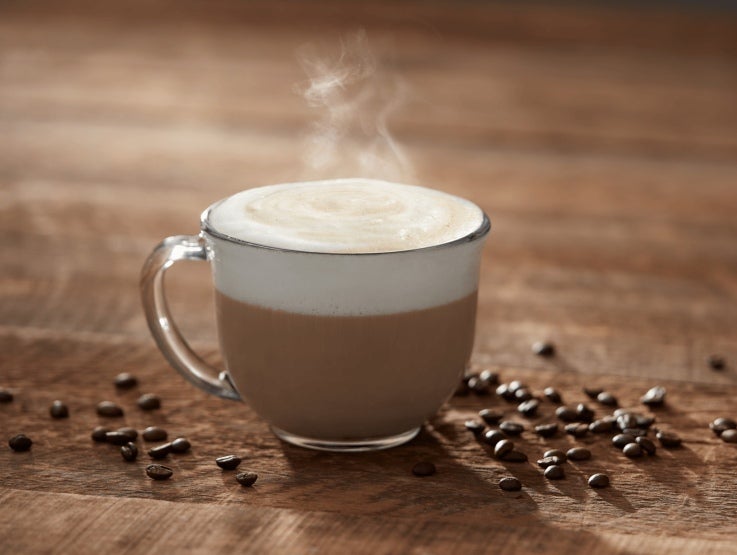 Taza transparente de cappuccino humeante rodeada de granos de café sobre una mesa de madera