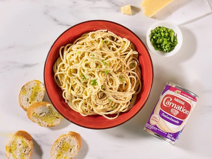 Tazón de espagueti con salsa blanca y especias, al lado una lata de CARNATION®, un tazón de cebollines y tostadas con aceite