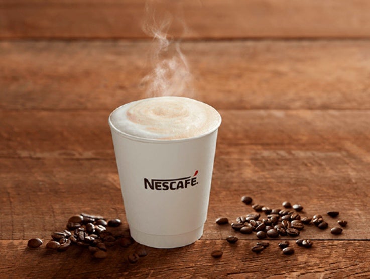 Vaso desechable blanco con el logo de NESCAFÉ® con una bebida caliente y alrededor granos de café tostados