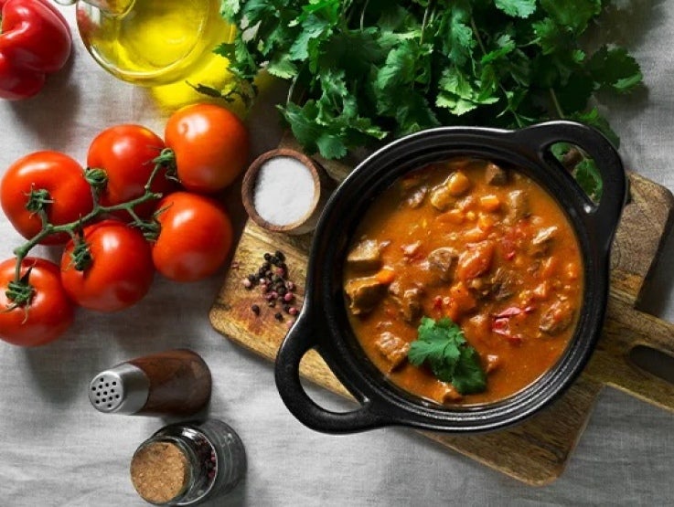 Olla de estofado rodeada por tomates, aceite, hierbas aromáticas y otros alimentos o productos culinarios