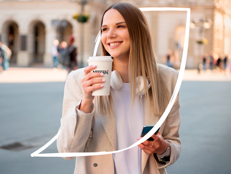 Mujer sonriente con auriculares alrededor del cuello, sosteniendo un vaso de café NESCAFÉ en una mano y un móvil en la otra