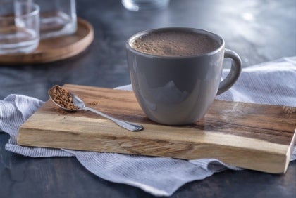 Una taza gris de chocolate caliente para máquinas, sobre una tabla de madera, con una cucharada de cacao en polvo.