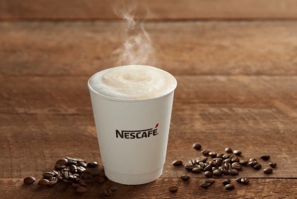 Taza humeante de NESCAFÉ® reposa entre granos de café para máquina esparcidos sobre una mesa