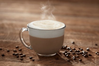 Taza transparente de cappuccino humeante rodeada de granos de café sobre una mesa de madera