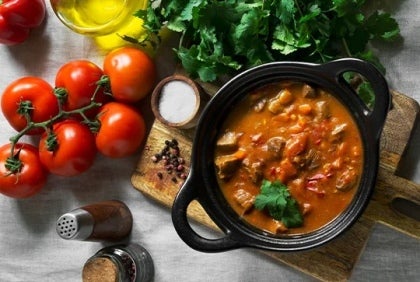 Olla de estofado rodeada por tomates, aceite, hierbas aromáticas y otros alimentos o productos culinarios