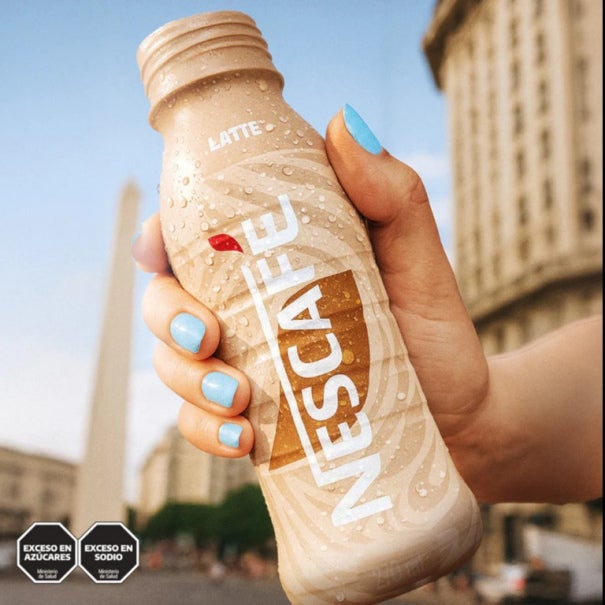 Mano sosteniendo una botella fría de NESCAFÉ Latte con gotas de agua, con fondo urbano desenfocado y edificios altos