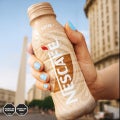 Mano sosteniendo una botella fría de NESCAFÉ Latte con gotas de agua, con fondo urbano desenfocado y edificios altos