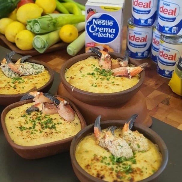 "3 platos de Chupe de jaibas junto a una caja de Crema de leche Nestlé, 5 tarros de Leche Evaporada Ideal y limones y puerros