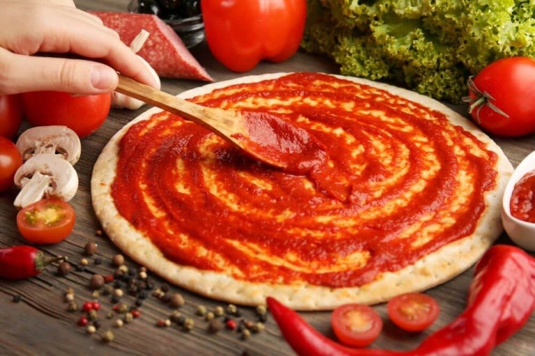 Mano con una cuchara de madera aplicando salsa de tomate sobre masa para pizza