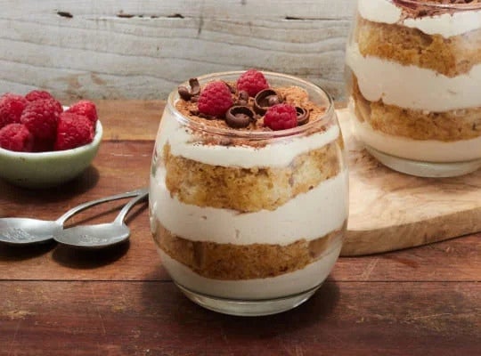 Dos vasos rellenos con un postre de tiramisú y decorado con chocolate y frambuesas