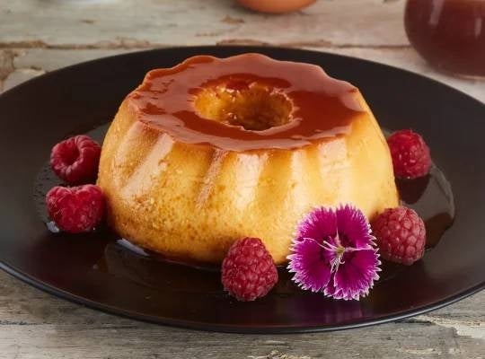 Crema Volteada Gourmet servida en un plato negro y decorada con NESTLÉ® Leche Condensada, flores y frambuesas
