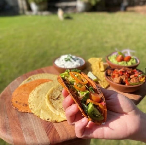 Mano sostiene un taco elaborado con tortillas de maíz y vegetales, al fondo los ingredientes