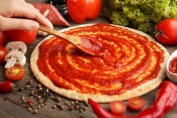 Mano con una cuchara de madera aplicando salsa de tomate sobre masa para pizza