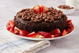 Pastel de chocolate cubierto con racimos de chocolate, rodeado de fresas en rodajas en un plato blanco.