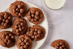 Un plato de galletas de chocolate con nueces junto a un vaso de leche sobre una superficie clara.