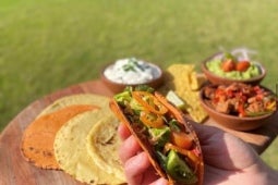 Mano sostiene un taco elaborado con tortillas de maíz y vegetales, al fondo los ingredientes