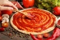 Mano con una cuchara de madera aplicando salsa de tomate sobre masa para pizza