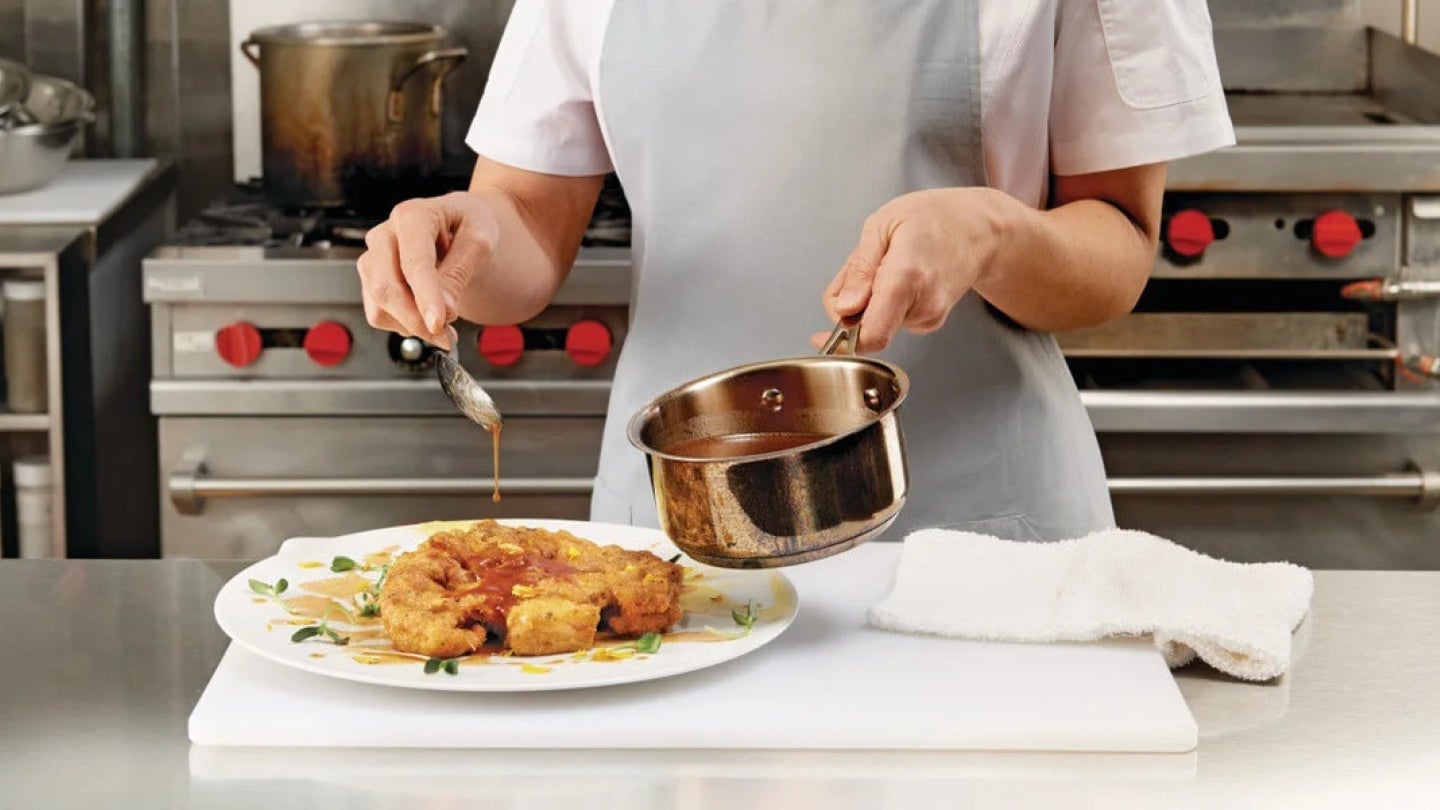 En una cocina comercial, una chef sostiene con una mano una olla y con la otra vierte una salsa con una cuchara a un platillo