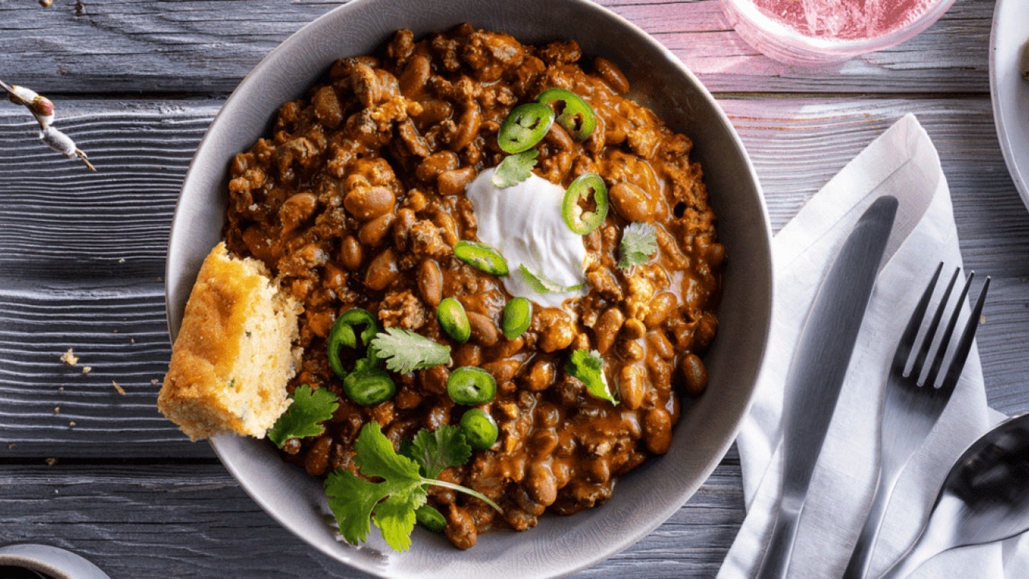 Vista cenital a un plato de chili con carne, cebollines, hierbas y un trozo de pan. Al lado de este un par de cubiertos sobre