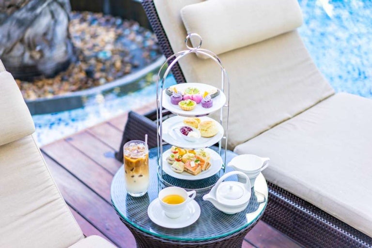 Juego de té con café con leche, té caliente y bocadillo en una mesa cerca de sillas junto a una piscina de hotel