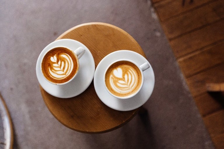 Dos tazas de café latte con arte en espuma, colocadas sobre una mesa de madera en un entorno acogedor.