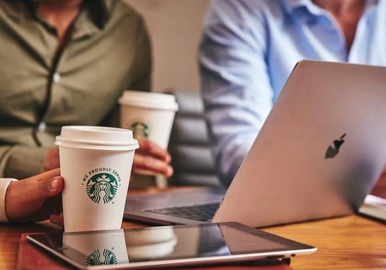 Personas en reunión de oficina compartiendo café para llevar mientras trabajan con laptops.