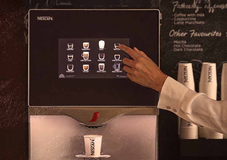 Mano seleccionando bebida en pantalla táctil de máquina de café con vaso debajo