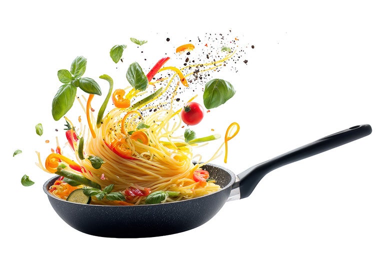  Pasta en sartén con ingredientes como tomates y hojas verdes en movimiento sobre fondo claro