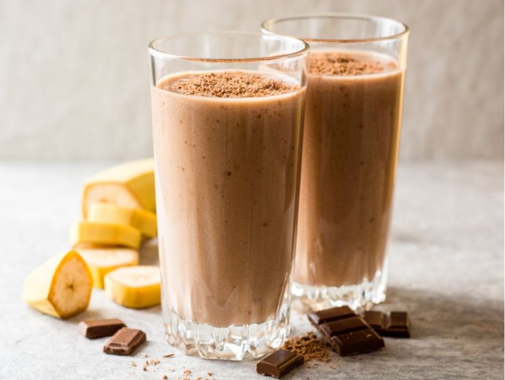 Vasos altos con batido cremoso de chocolate y plátano decorados con cacao junto a fruta fresca.