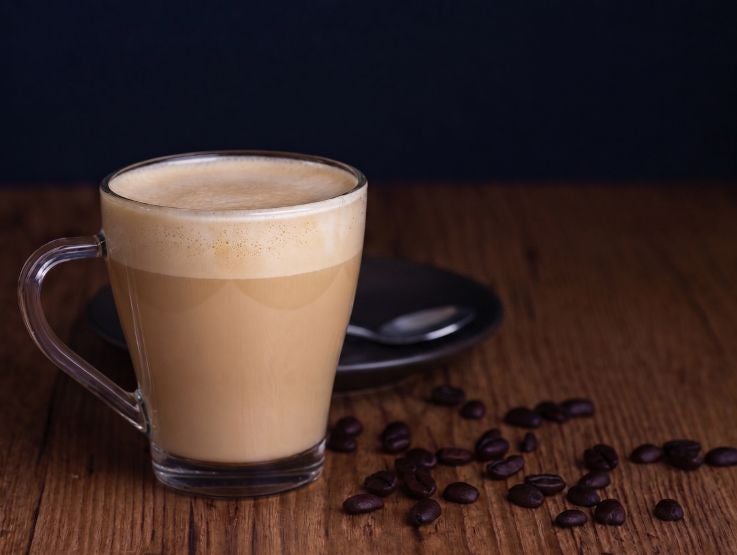 Taza de cristal con cappuccino espumoso sobre mesa de madera rodeada de granos de café.