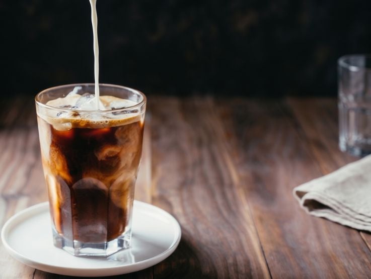 Vaso de cristal con café helado con hielo mientras se vierte un chorro de crema desde arriba.