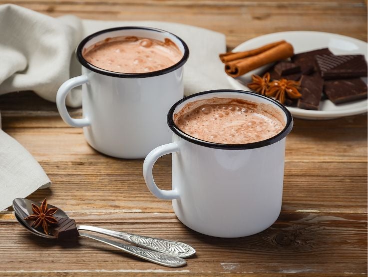 Tazas de peltre blanco con chocolate caliente espumoso sobre mesa de madera rústica con canela.