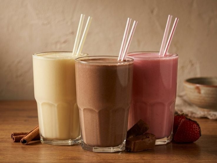 Tres vasos con batidos de vainilla, chocolate y fresa acompañados de sus ingredientes frescos.