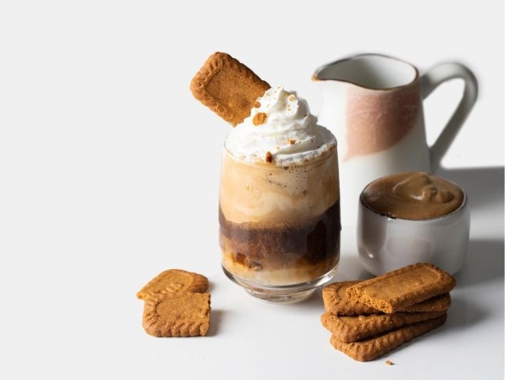 Vaso de café frío cubierto con crema batida y una galleta crujiente sobre mesa de color blanco.