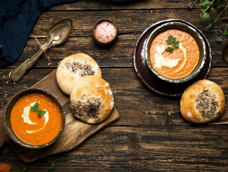 Dos tazones de sopa cremosa de tomate acompañados de panes artesanales sobre mesa de madera.