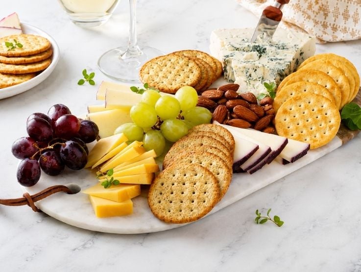 Tabla de mármol con galletas saladas, variedad de quesos, uvas frescas y almendras tostadas.